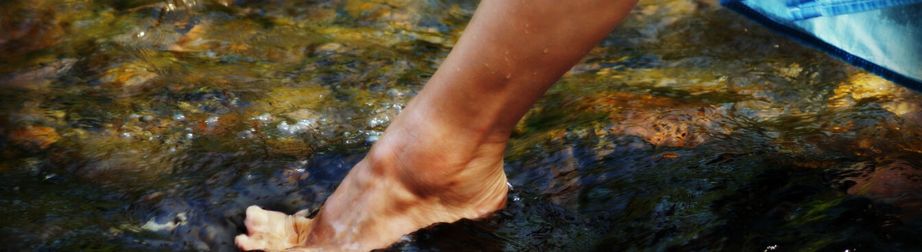 Eine Person watet mit nackten Füßen durch ein flaches, fließendes Gewässer. Die Jeans sind leicht hochgekrempelt, um nicht nass zu werden. Die Szene wirkt erfrischend und naturverbunden. Eine Person watet mit nackten Füßen durch ein flaches, fließendes Gewässer. Die Jeans sind leicht hochgekrempelt, um nicht nass zu werden. Die Szene wirkt erfrischend und naturverbunden.