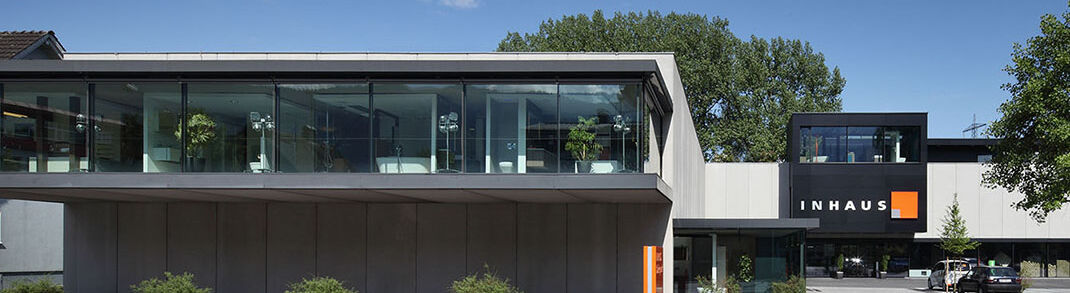Bäderpark Hohenems. Die Badausstellung in Vorarlberg. Ein modernes, zweistöckiges Gebäude mit großen Glasfenstern. Auf der Fassade ist "INHAUS" zu lesen. Vor dem Gebäude gibt es eine Straße und einige Büsche.
