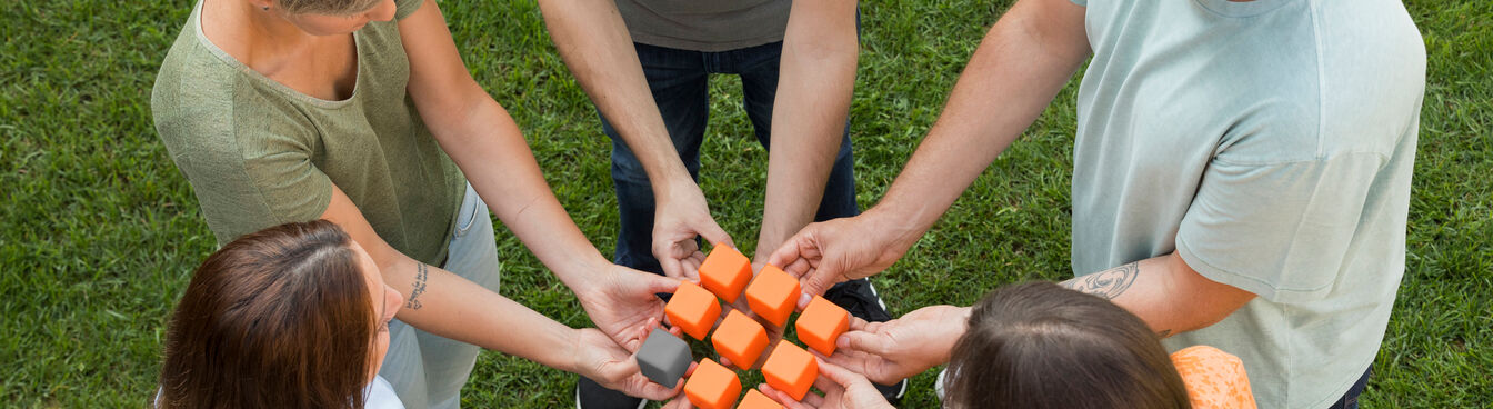 Fünf Personen stehen im Kreis auf einer Wiese und halten gemeinsam orangefarbene Würfel in der Mitte. Sie scheinen an einer gemeinsamen Aktivität teilzunehmen. Fünf Personen stehen im Kreis auf einer Wiese und halten gemeinsam orangefarbene Würfel in der Mitte. Sie scheinen an einer gemeinsamen Aktivität teilzunehmen.