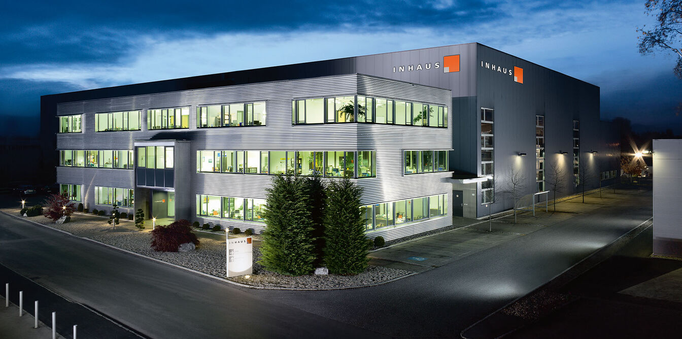 Ein modernes Bürogebäude bei Nacht, beleuchtet von innen. Es hat drei Etagen, große Fenster und eine gepflegte Umgebung mit Bäumen und Gehwegen. Ein modernes Bürogebäude bei Nacht, beleuchtet von innen. Es hat drei Etagen, große Fenster und eine gepflegte Umgebung mit Bäumen und Gehwegen.