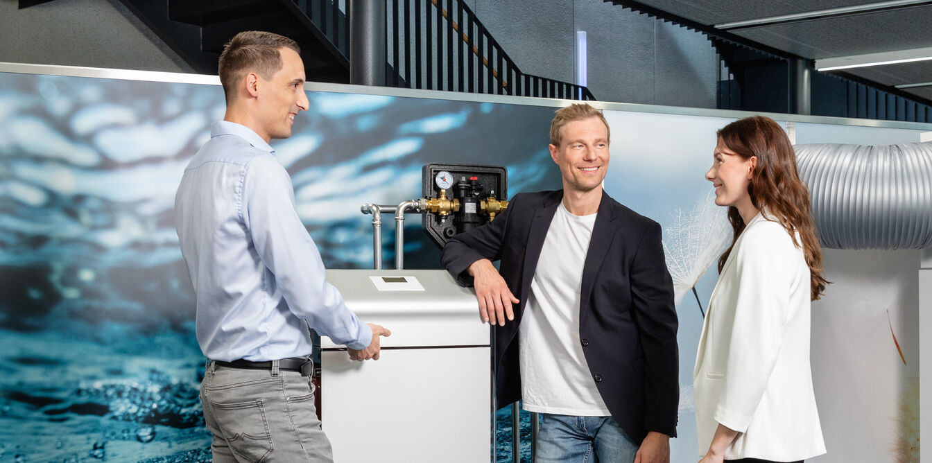 Eine Gruppe von drei Personen steht l&auml;chelnd um ein technisches Ger&auml;t in einem modernen Energiepark, vor einem Hintergrund mit Wasserbild.