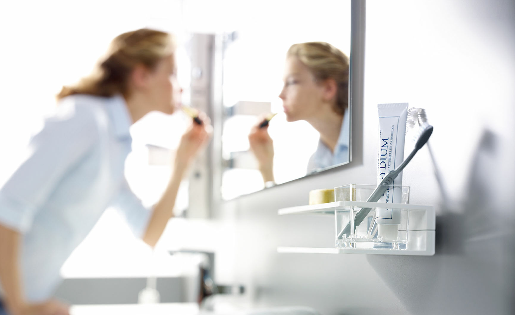 Eine Person schminkt sich vor einem Spiegel. Im Vordergrund ist ein Zahnpasta- und Zahnbürstenhalter an der Wand montiert. Eine Person schminkt sich vor einem Spiegel. Im Vordergrund ist ein Zahnpasta- und Zahnbürstenhalter an der Wand montiert.