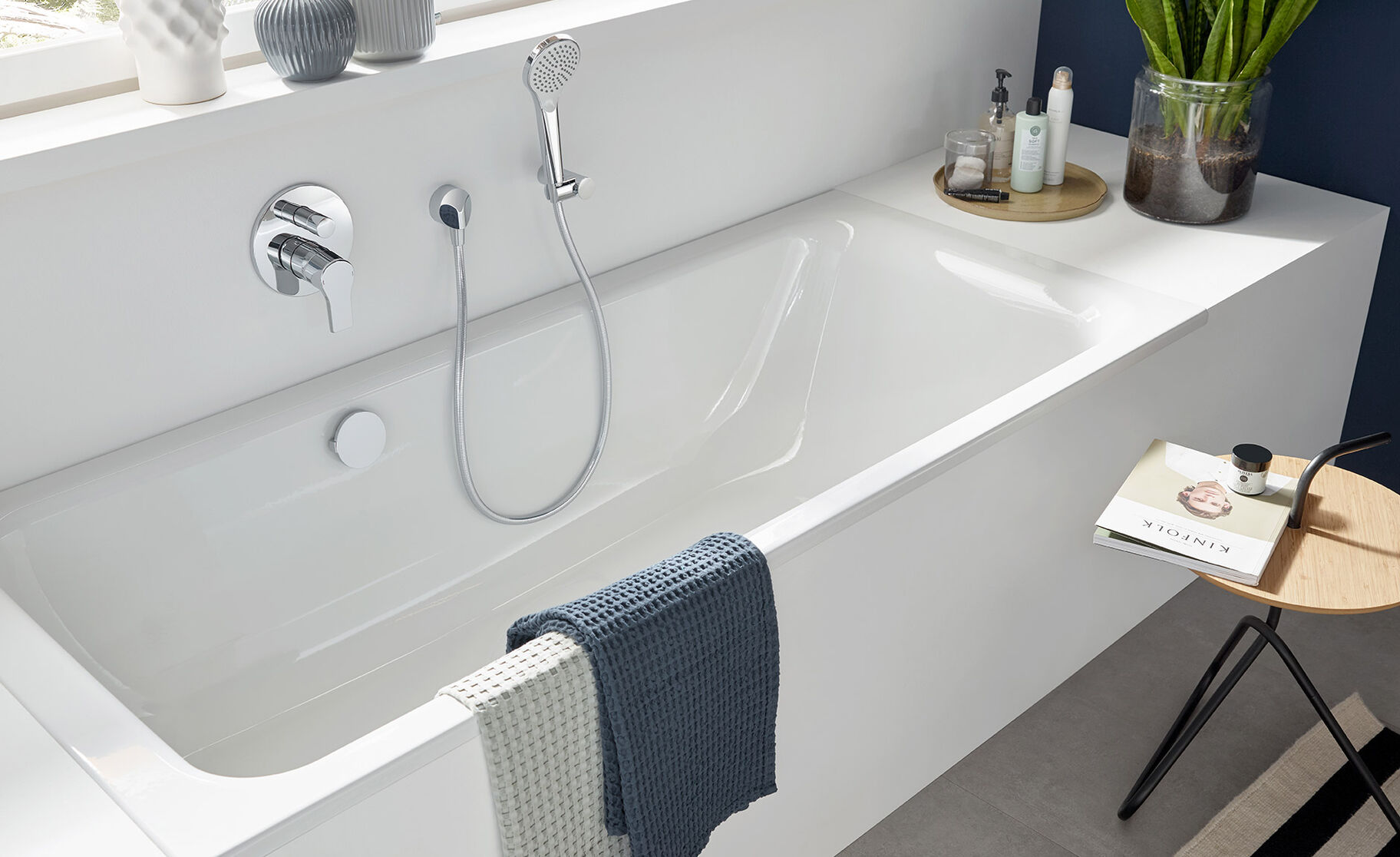 Modernes Bad mit weißer Badewanne, Pflanzen, dekorativen Vasen und gestreiftem Teppich. Viel natürliches Licht und schlichte Eleganz prägen den Raum. Modernes Bad mit weißer Badewanne, Pflanzen, dekorativen Vasen und gestreiftem Teppich. Viel natürliches Licht und schlichte Eleganz prägen den Raum.