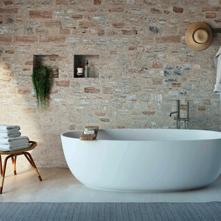 Freistehende wei&szlig;e Badewanne vor einer rustikalen Steinwand. Handt&uuml;cher auf einem Hocker, daneben ein Bademantel und ein Hut an der Wand. Pflanze in einem Regal.