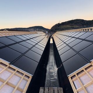 Solarpaneele auf einem V-f&ouml;rmigen Dach, eingebettet in eine h&uuml;gelige Landschaft bei Sonnenuntergang.