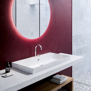 Modernes Badezimmer mit roter Wand, rundem Spiegel mit Beleuchtung, wei&szlig;em Waschbecken und Holzbank auf gemustertem Boden.