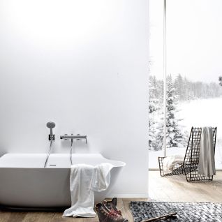 Moderne Badewanne vor einer schlichten wei&szlig;en Wand, mit Ausblick auf verschneite Landschaft. Holzfu&szlig;boden, gem&uuml;tliches Ambiente mit Handtuch und Winterstiefeln.