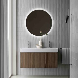 Modernes Bad mit Holz-Waschtisch, rundem beleuchtetem Spiegel und grauen Fliesen. Eine flauschige Badematte und minimalistische Deko erg&auml;nzen das Design.