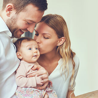 Ein Paar hält liebevoll ein Baby. Die Mutter gibt dem Baby einen Kuss, während der Vater es hält. Es ist eine warme und liebevolle Familienszene.