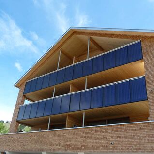 Ein modernes Geb&auml;ude mit Solarpaneelen an den Balkonen, gebaut aus Ziegel und Holz, unter einem blauen Himmel.