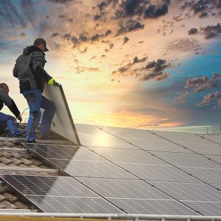 Zwei Arbeiter installieren Solarpaneele auf einem Ziegeldach, w&auml;hrend die Sonne am Horizont untergeht und den Himmel in warmen Farben erleuchtet.