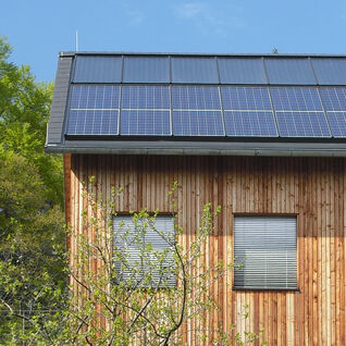 Ein modernes Holzhaus mit Solarpanels auf dem Dach, umgeben von grüner Natur und einem Baum, unter einem blauen Himmel mit wenigen Wolken. Ein modernes Holzhaus mit Solarpanels auf dem Dach, umgeben von grüner Natur und einem Baum, unter einem blauen Himmel mit wenigen Wolken.