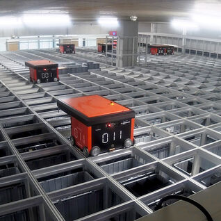 Ein automatisiertes Lagersystem mit mehreren orangefarbenen Robotern, die auf einem Gitterrahmen fahren, um Waren effizient zu bewegen.