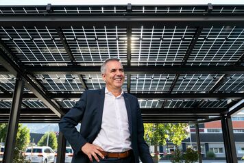 Ein Mann in einem Anzug steht unter einem Solardach, das aus mehreren Solarpaneelen besteht. Er l&auml;chelt und hat eine Hand in der Hosentasche.