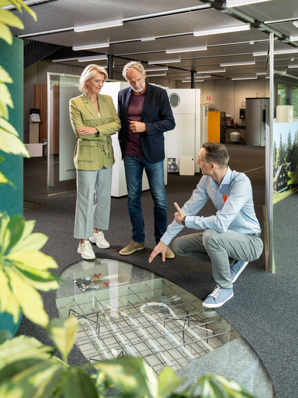 Drei Personen befinden sich in einem modernen Raum. Ein Mann kniet und zeigt auf den Boden, während das Paar interessiert zusieht. Pflanzen sind im Vordergrund. Drei Personen befinden sich in einem modernen Raum. Ein Mann kniet und zeigt auf den Boden, während das Paar interessiert zusieht. Pflanzen sind im Vordergrund.