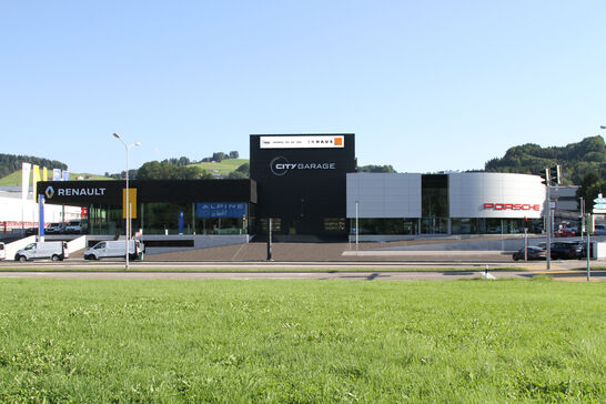 Ein modernes Gebäude mit den Marken Inhaus, Renault und Porsche, umgeben von grünen Hügeln und einem Parkplatz im Vordergrund. Ein modernes Gebäude mit den Marken Inhaus, Renault und Porsche, umgeben von grünen Hügeln und einem Parkplatz im Vordergrund.