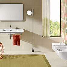 Ein stilvolles Badezimmer mit einem Waschtisch und Spiegel links, WC und Bidet rechts. Eine bunte Akzentwand und ein Hocker verleihen dem Raum Farbe.