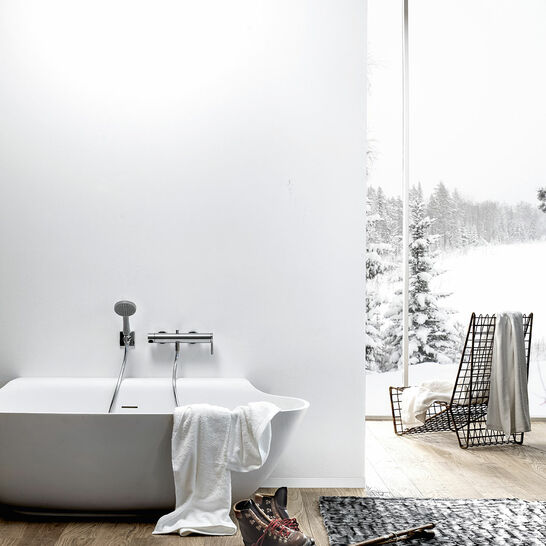 Moderne Badewanne vor einer schlichten weißen Wand, mit Ausblick auf verschneite Landschaft. Holzfußboden, gemütliches Ambiente mit Handtuch und Winterstiefeln. Moderne Badewanne vor einer schlichten weißen Wand, mit Ausblick auf verschneite Landschaft. Holzfußboden, gemütliches Ambiente mit Handtuch und Winterstiefeln.