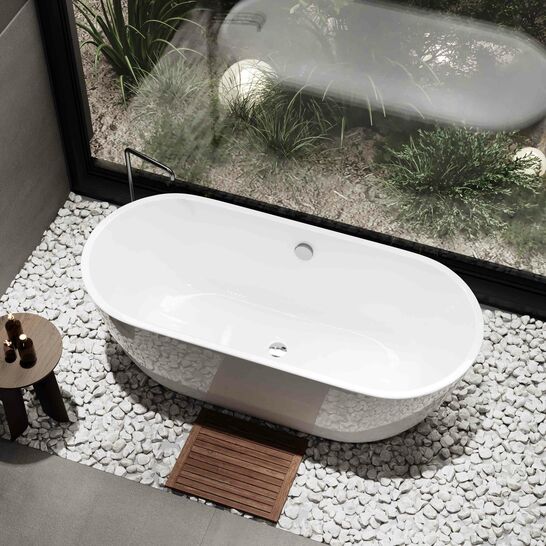 Freistehende Badewanne auf wei&szlig;en Kieselsteinen, mit h&ouml;lzernem Badvorleger und kleinem Beistelltisch. Gro&szlig;es Fenster bietet Blick auf Garten.