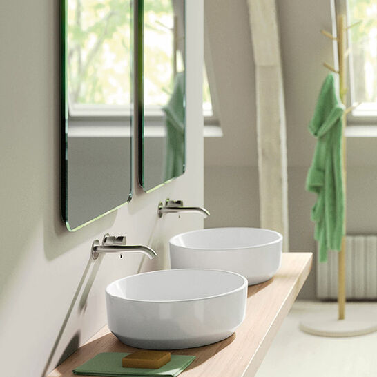Ein stilvolles Badezimmer mit zwei runden Aufsatzwaschbecken auf einem Holzbrett. Dar&uuml;ber befinden sich zwei Spiegel. Im Hintergrund h&auml;ngt ein gr&uuml;nes Handtuch. Modern und elegant gestaltet.
