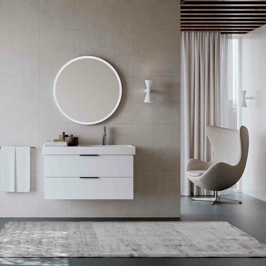 Modernes Bad mit rundem Spiegel, wei&szlig;em Waschtisch, grauem Teppich, eleganten M&ouml;beln und nat&uuml;rlichem Licht durch gro&szlig;es Fenster.