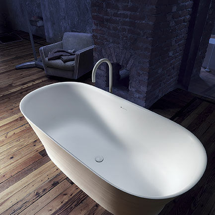Freistehende Badewanne in einem rustikalen Raum mit Holzboden und Backsteinwand, kombiniert mit einem gem&uuml;tlichen Sessel im Hintergrund.