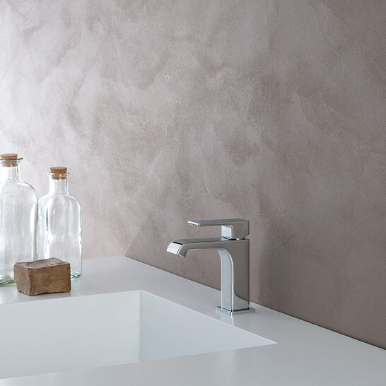 Modernes Waschbecken mit eckigem Wasserhahn auf weißer Fläche. Links stehen zwei Glasflaschen und ein Stück Seife. Die Wand hat eine beige Textur. Modernes Waschbecken mit eckigem Wasserhahn auf weißer Fläche. Links stehen zwei Glasflaschen und ein Stück Seife. Die Wand hat eine beige Textur.