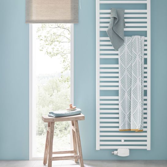 Wei&szlig;er Handtuchheizk&ouml;rper an blauer Wand, dekoriert mit zwei Handt&uuml;chern. Neben einem Fenster steht ein Holzhocker mit gefalteten Handt&uuml;chern.