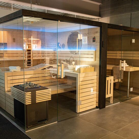 Ein moderner Saunabereich mit Glaswänden. Die Inneneinrichtung besteht aus Holz mit Sitzbänken, und es gibt eine Saunasteuerung. Die Beleuchtung schafft eine entspannende Atmosphäre, und der Raum wirkt sauber und gut organisiert. Inhaus Baederpark Hohenems Sauna