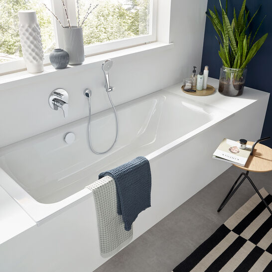 Modernes Bad mit weißer Badewanne, Pflanzen, dekorativen Vasen und gestreiftem Teppich. Viel natürliches Licht und schlichte Eleganz prägen den Raum.
