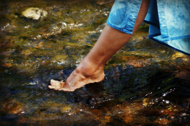 Eine Person watet mit nackten Füßen durch ein flaches, fließendes Gewässer. Die Jeans sind leicht hochgekrempelt, um nicht nass zu werden. Die Szene wirkt erfrischend und naturverbunden. Eine Person watet mit nackten Füßen durch ein flaches, fließendes Gewässer. Die Jeans sind leicht hochgekrempelt, um nicht nass zu werden. Die Szene wirkt erfrischend und naturverbunden.