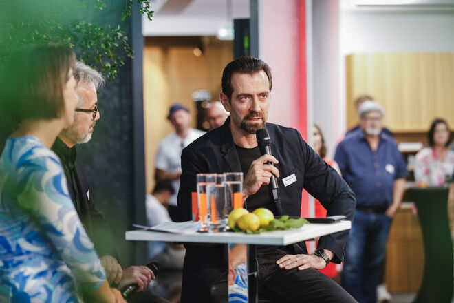ictor Vasilev h&auml;lt ein Mikrofon und spricht bei einer Veranstaltung. Er sitzt an einem Tisch mit Gl&auml;sern und Zitronen darauf. Um ihn herum stehen weitere Personen und h&ouml;ren zu.