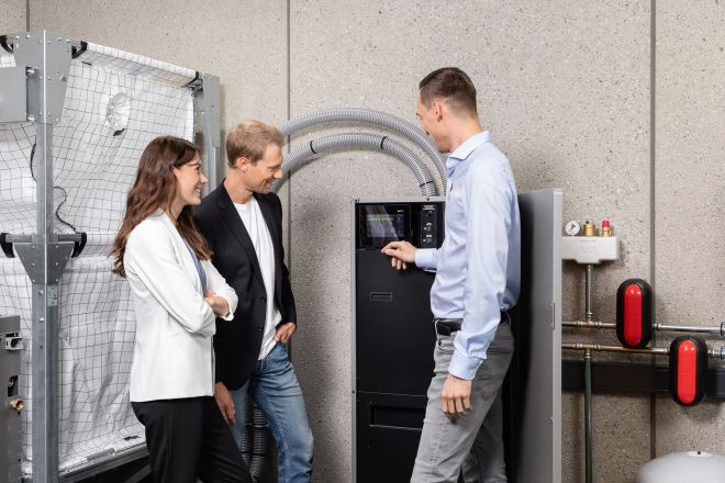 Drei Personen stehen in einem technischen Raum. Ein Mann in einem blauen Hemd erkl&auml;rt etwas an einem gro&szlig;en Ger&auml;t mit Display, w&auml;hrend ein Paar interessiert zuh&ouml;rt. 