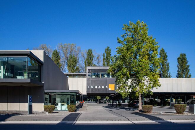 Ein modernes Geb&auml;ude mit der Aufschrift "INHAUS". Es hat eine klare, geradlinige Architektur mit gro&szlig;en Fenstern. Ein gro&szlig;er Baum steht auf dem Vorplatz, der von Pflanzen ges&auml;umt ist.
