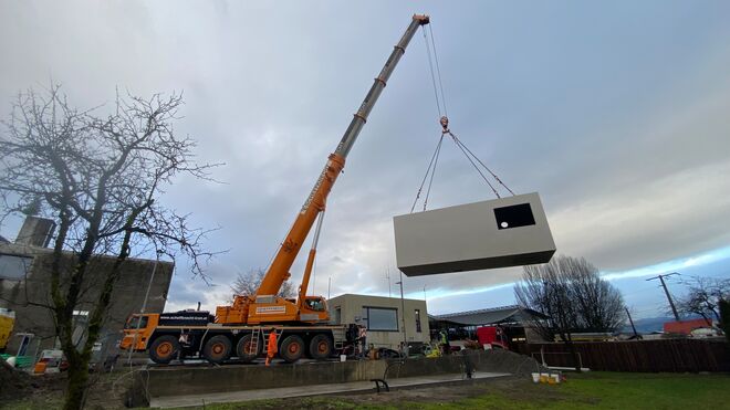 Ein gro&szlig;er Kran hebt ein vorgefertigtes Geb&auml;udetei in Form eines Containers an. Das Modul hat ein Fenster. Die Szene spielt im Freien.
