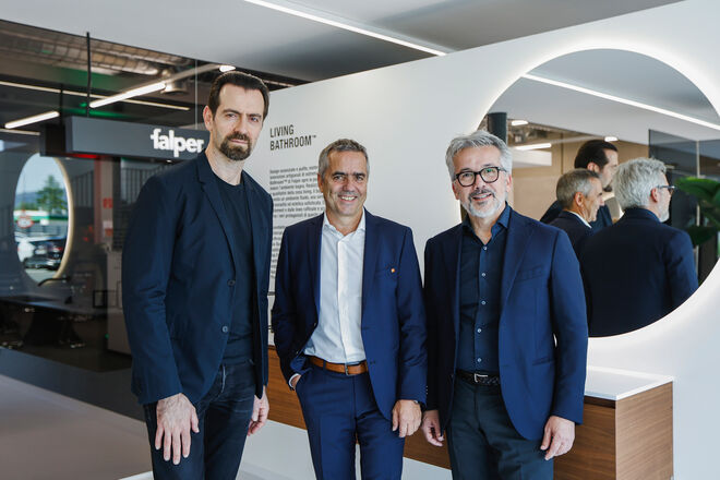 Drei M&auml;nner in Anz&uuml;gen stehen vor einem modernen Ausstellungsraum mit einem runden Spiegel und der Aufschrift "LIVING BATHROOM&trade;" im Hintergrund.