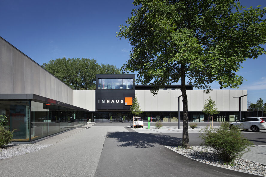Ein modernes Gebäude mit der Aufschrift "INHAUS" auf der Fassade. Der Außenbereich ist gepflegt mit Bäumen und Gehwegen unter einem klaren blauen Himmel. Ein modernes Gebäude mit der Aufschrift "INHAUS" auf der Fassade. Der Außenbereich ist gepflegt mit Bäumen und Gehwegen unter einem klaren blauen Himmel.