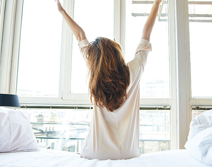 Eine Person sitzt auf einem Bett und streckt sich vor einem großen, lichtdurchfluteten Fenster. Der Raum wirkt hell und einladend. Eine Person sitzt auf einem Bett und streckt sich vor einem großen, lichtdurchfluteten Fenster. Der Raum wirkt hell und einladend.